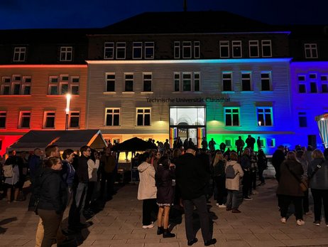 Hier ist das mit Regenbogenfarben angestrahlte TU Hauptgebäude mit einer Menschenmenge davor zu sehen.