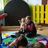 Ein Junge im Teenager-Alter sitzt mit einer Puppe in der Hand auf einem farbigen Teppich und spielt mit kleinen Kindern.